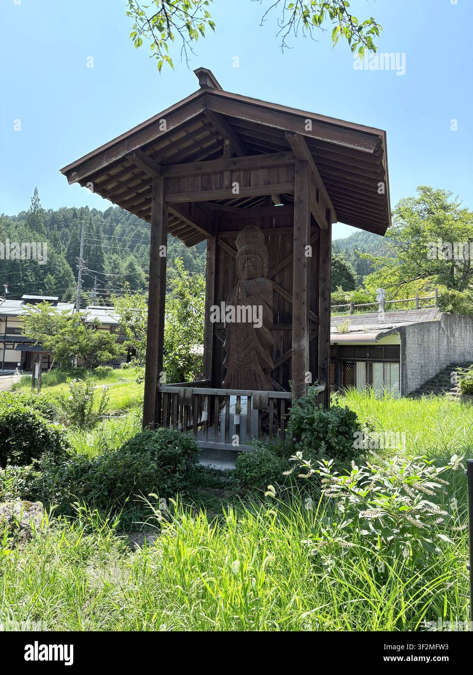 30 agosto 2025, Takayama, Giappone – Statua religiosa in legno consumata protetta da un tradizionale baldacchino in legno scuro circondata da vegetazione verde rigogliosa nella storica Takayama. - Immagine stock catturata con smartphone