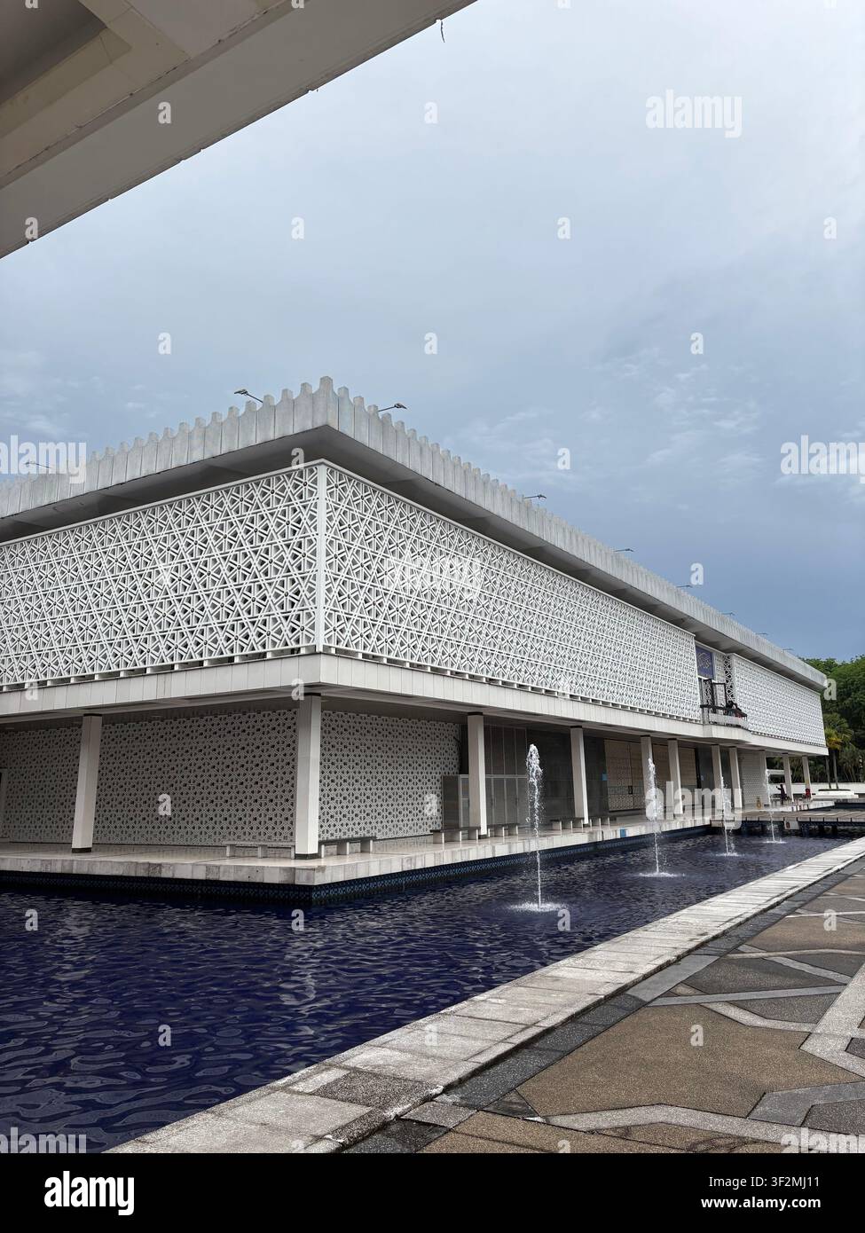 4 maggio 2025, Kuala Lumpur, Malesia – Vista drammatica a bassa angolazione della facciata geometrica bianca a griglia della moschea nazionale Masjid Negara con una piscina fontana riflettente. - Immagine stock catturata con smartphone