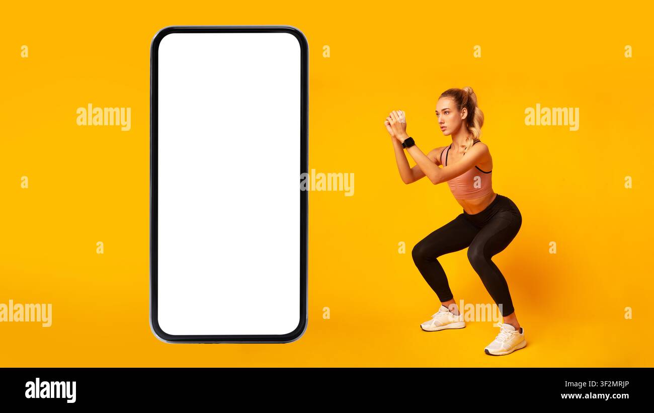 Modella femminile che esegue un esercizio di fitness con squat e mockup di smartphone, sfondo giallo Foto Stock