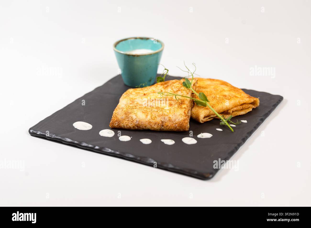 Scatto ad angolo alto in studio di pancake dorati piegati serviti con panna acida in una tazza turchese, microverdure fresche Foto Stock