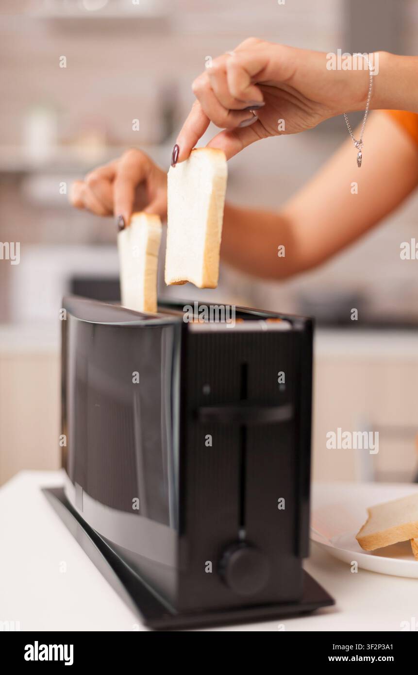 Donna d'affari mangia pane tostato mentre lavora su un laptop durante la colazione, multitasking in cucina prima della giornata in ufficio, evidenziando la preparazione del cibo, la routine domestica e lo stile di vita quotidiano. Foto Stock