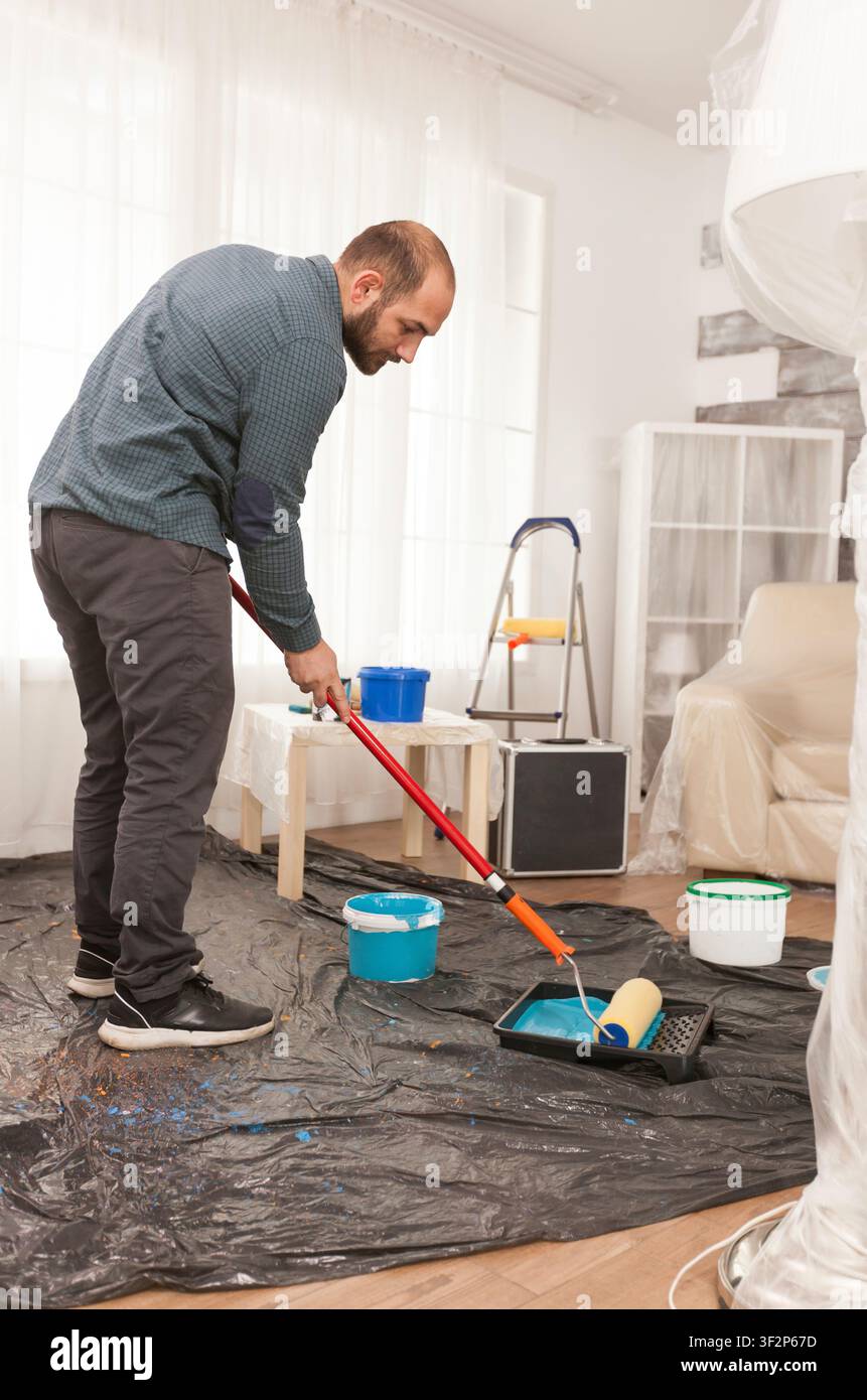 Persona che tiene un rullo per la pittura per un progetto murale mentre strumenti per la pittura, coperture protettive e preparazione del muro modellano un compito pratico di miglioramento della casa. Foto Stock
