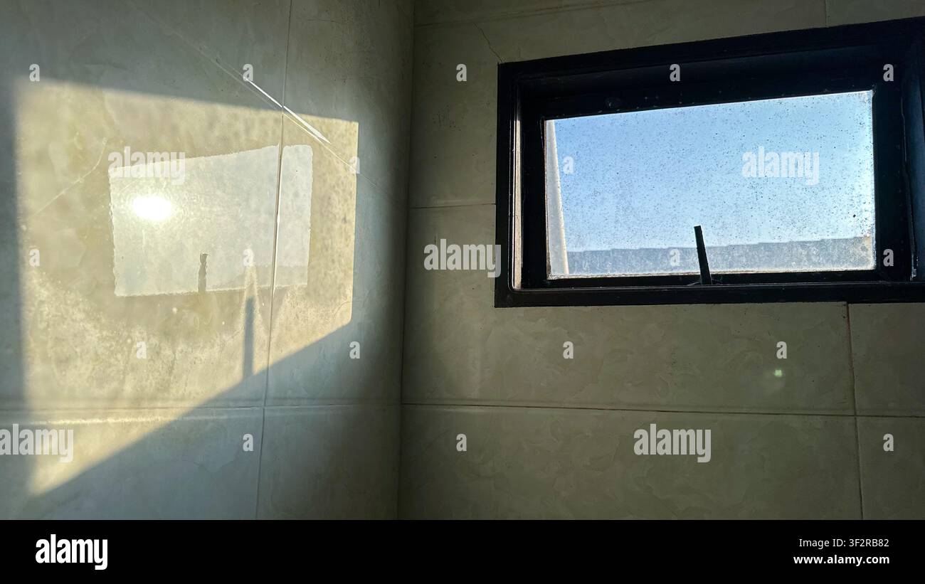 La luce del sole che proietta riflessi di ombre geometriche luminose su una parete piastrellata attraverso una piccola cornice quadrata della finestra all'interno di una stanza moderna a Johor Bahru, Malesia - Immagine stock catturata con smartphone