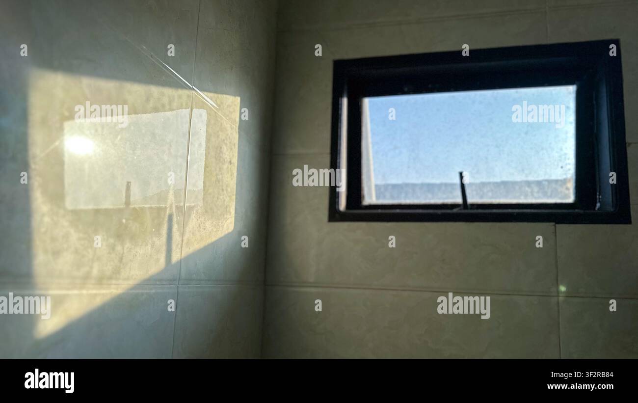 La luce del sole che proietta riflessi di ombre geometriche luminose su una parete piastrellata attraverso una piccola cornice quadrata della finestra all'interno di una stanza moderna a Johor Bahru, Malesia - Immagine stock catturata con smartphone