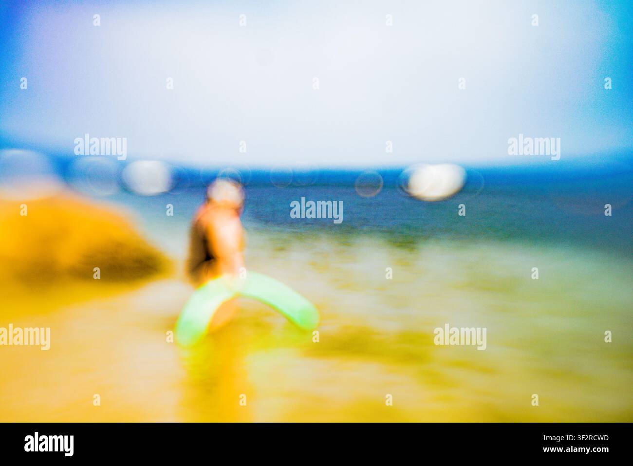Persona sfocata che tiene una canna da piscina verde in piedi su una spiaggia soleggiata al bordo dell'acqua Foto Stock