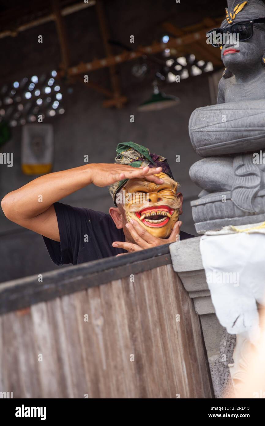 Un interprete che sbircia oltre una partizione di legno indossando una maschera balinese Bondres sorridente con bocca larga. Foto Stock