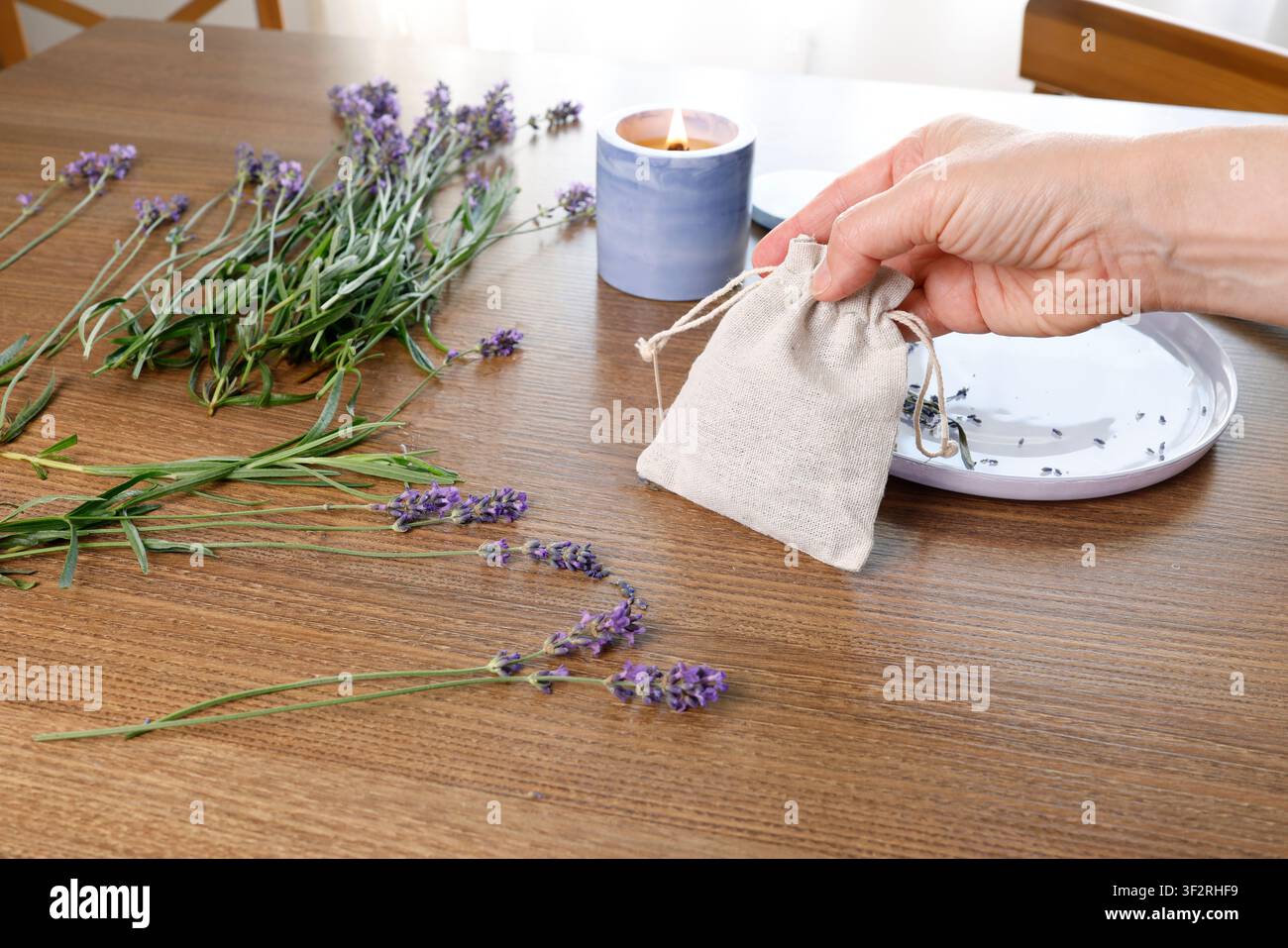Mano di donna che tiene un piccolo sacchetto di lino pieno di lavanda essiccata. Candela profumata che brucia su un tavolo di legno vicino a una composizione di bouquet di erbe. Fatto a mano Foto Stock