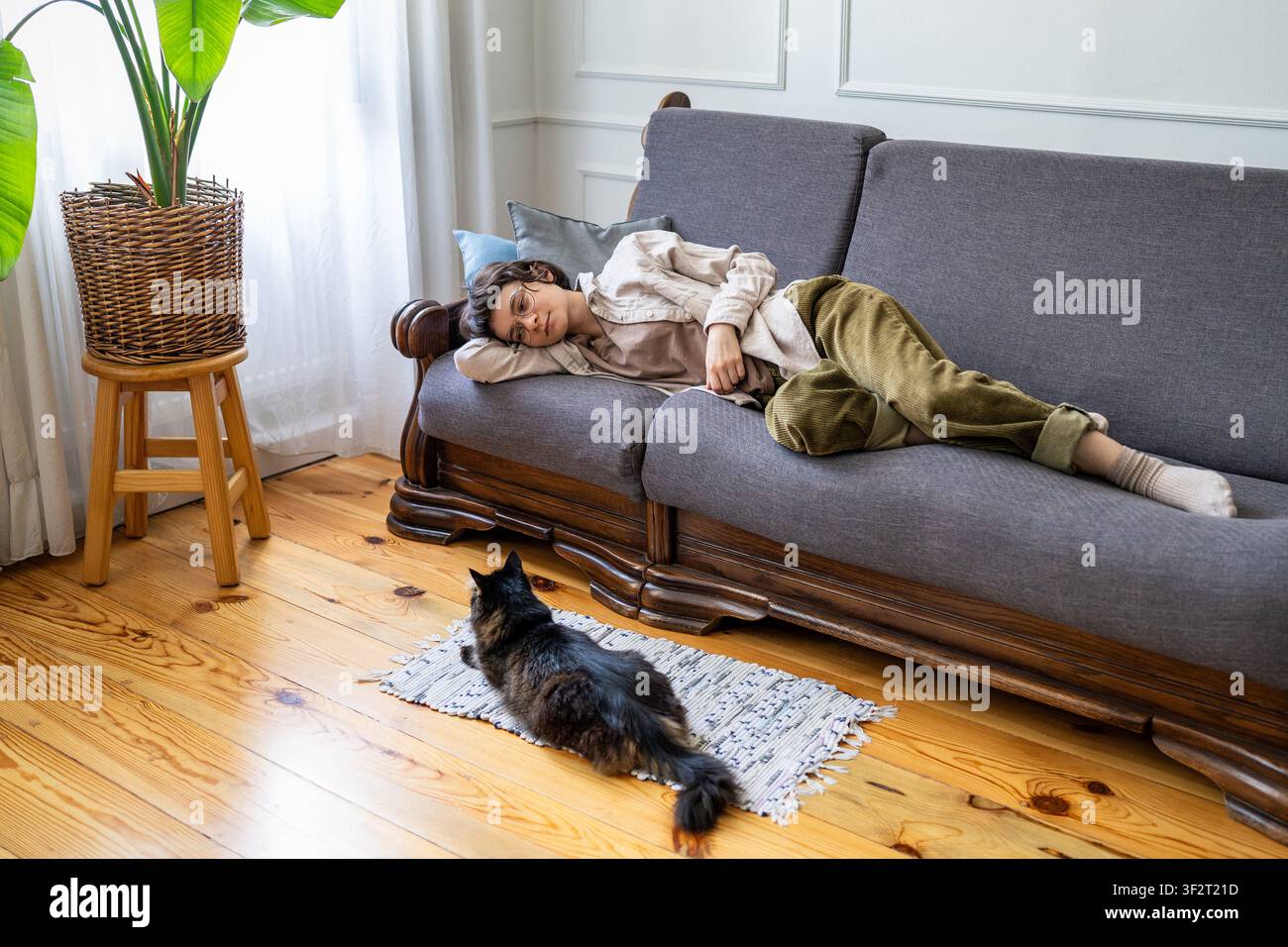 Donna pigra e felice che si rilassa pacificamente sul divano e osserva il gatto domestico che riposa sul tappeto nella stanza. Foto Stock