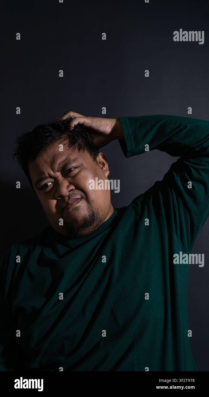 Ritratto umoristico ma cupo di un adulto indonesiano incerto che indossa una camicia verde scuro e si gratta la testa su uno sfondo nero profondo. Il Foto Stock