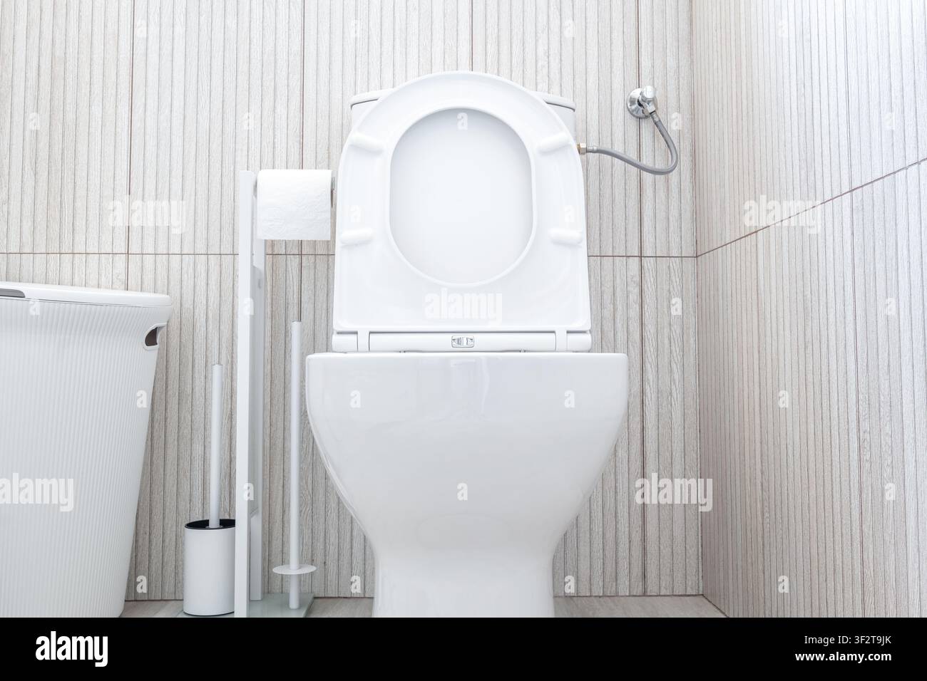 Vista frontale di una moderna tazza di toilette in ceramica bianca con seduta e coperchio aperti, portarotolo, spazzolino e piccolo cestino della spazzatura su uno sfondo di parete piastrellata luce Foto Stock