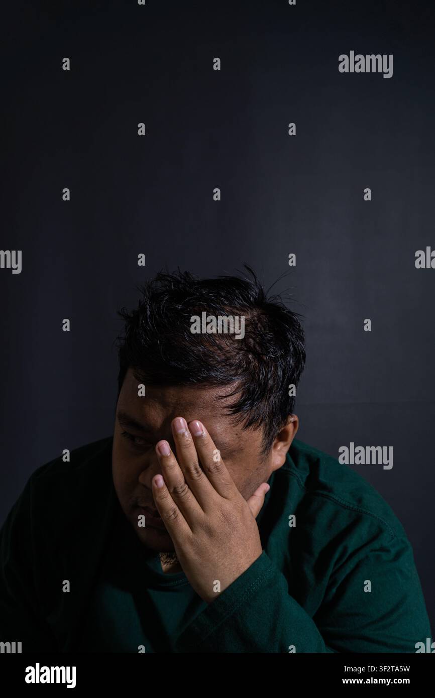 Ritratto malinconico di un uomo del sud-est asiatico che indossa una camicia verde scuro che copre un lato del viso con una mano su uno sfondo nero solido. Il sotto Foto Stock