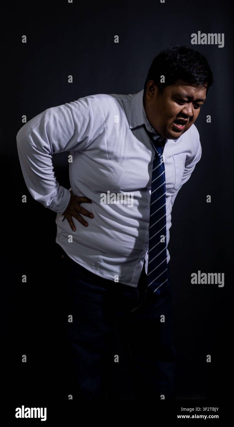 Uomo asiatico adulto affaticato che tiene la parte bassa della schiena dolorante, si appoggia all'indietro con un'espressione facciale smorfiosa su uno sfondo nero solido. L'ov Foto Stock