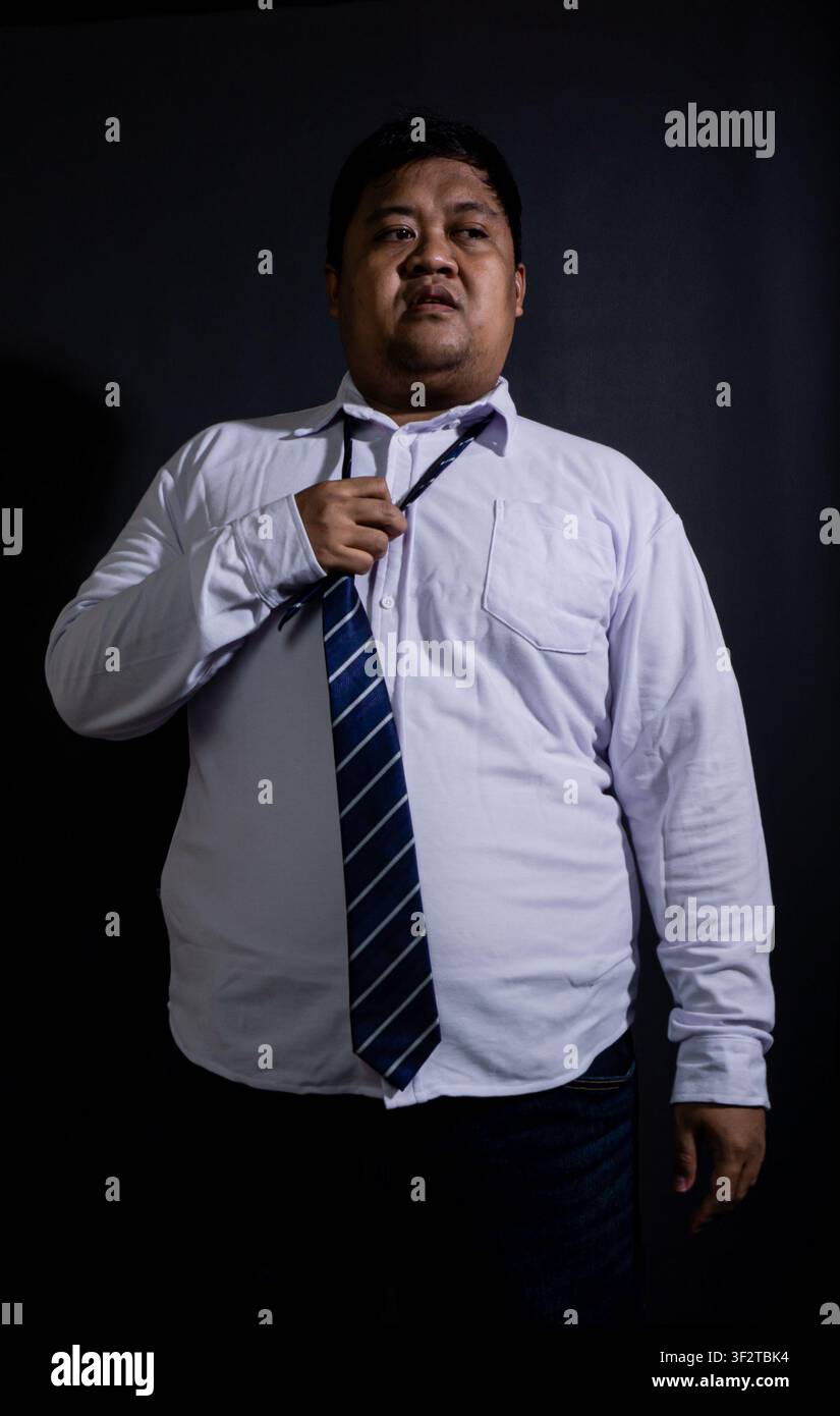 Uomo asiatico adulto stanco che allenta una cravatta blu a righe, cercando sollievo dopo una lunga giornata lavorativa su uno sfondo nero solido. L'uomo d'affari in sovrappeso Foto Stock