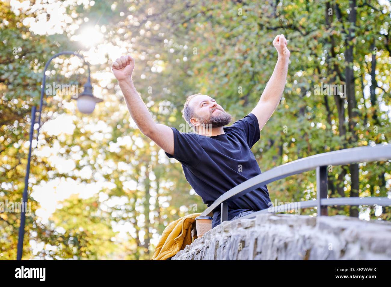 Uomo alza i pugni in segno di vittoria mentre è in piedi su un ponte del parco alla luce del sole autunnale. Scena all'aperto energetica per successo, realizzazione, motivazione, libertà o pubblicità lifestyle Foto Stock