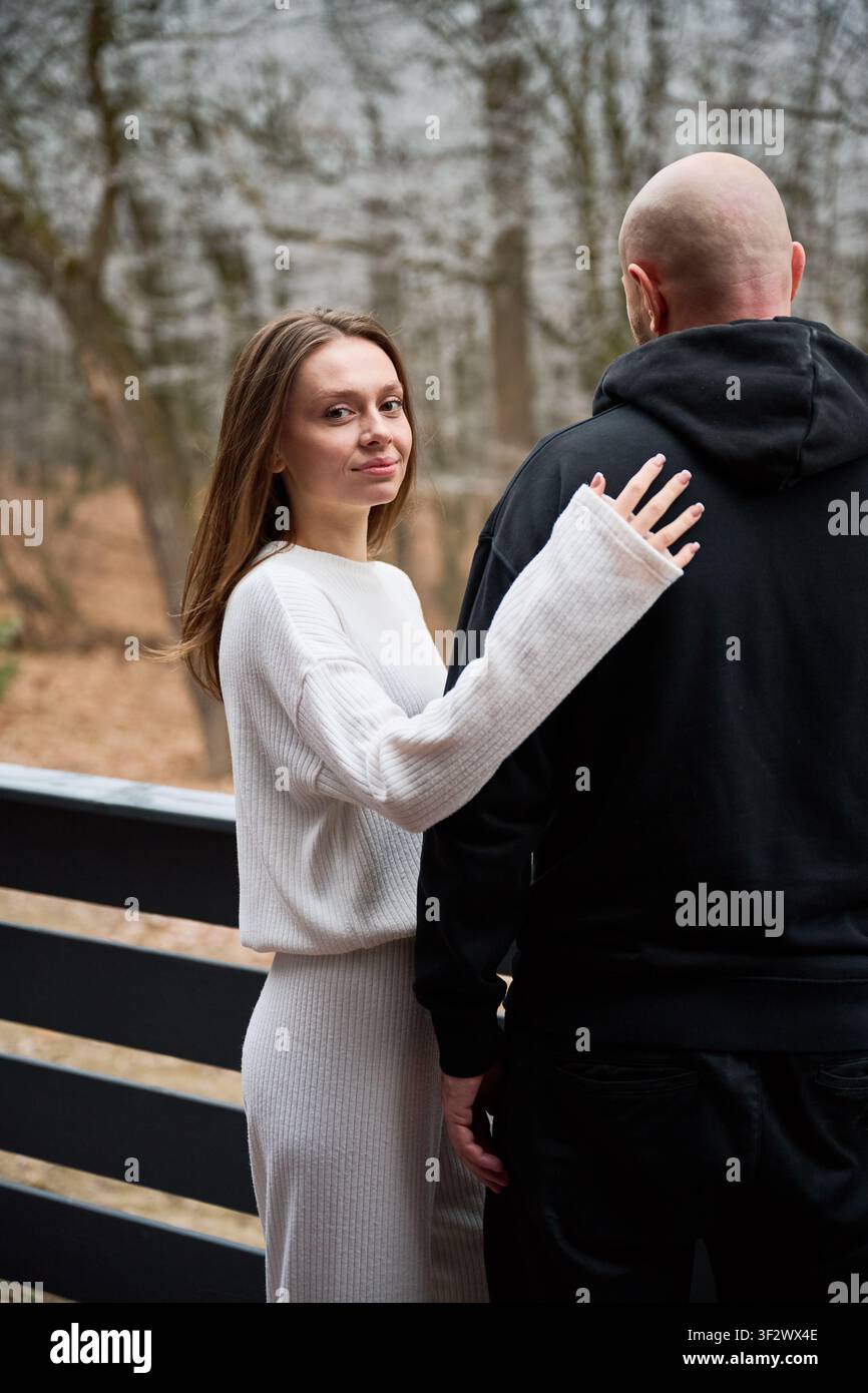 Coppia che gode di una scena tranquilla all'aperto sulla terrazza, donna che tocca la schiena dell'uomo con affetto, guardando la telecamera, esprimendo connessione, fiducia e uno stile di vita rilassato circondato da alberi Foto Stock