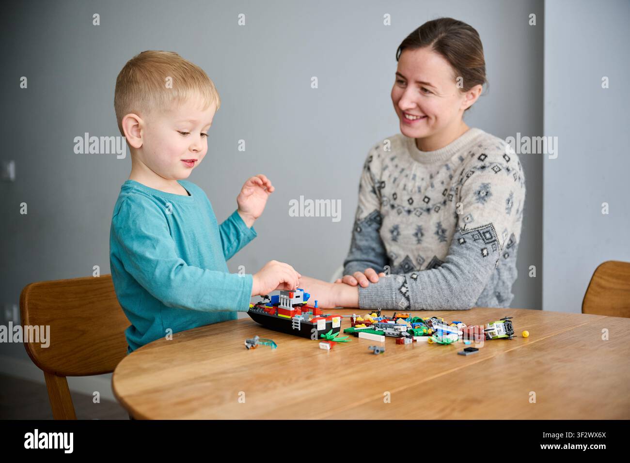 Ragazzo che gioca con blocchi di costruzione con la madre. Seduto al tavolo a casa, costruisce insieme un modello di giocattolo, genitore e bambino sorridenti, momento di legame familiare Foto Stock