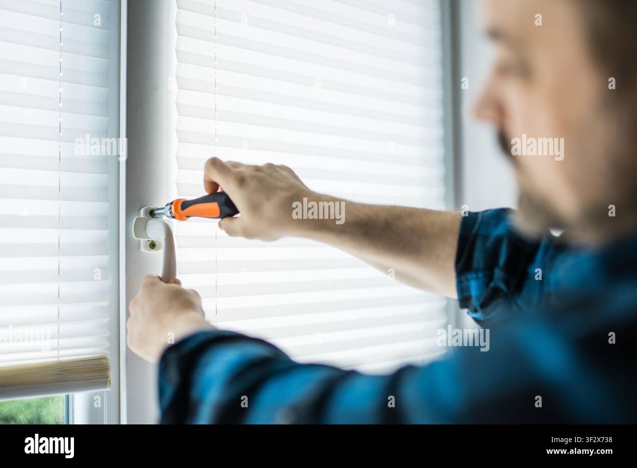 Uomo con una camicia a quadri blu che usa un cacciavite per riparare o regolare una maniglia di finestra in plastica bianca Foto Stock