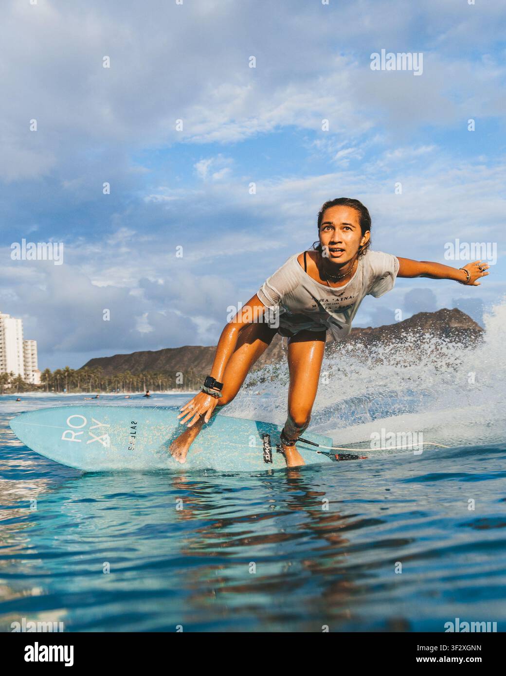 WAIKIKI, HONOLULU, HAWAII - 3 MAGGIO 2022: Una donna traccia una curva su una tavola blu con il cratere di Diamond Head alle sue spalle. Foto Stock
