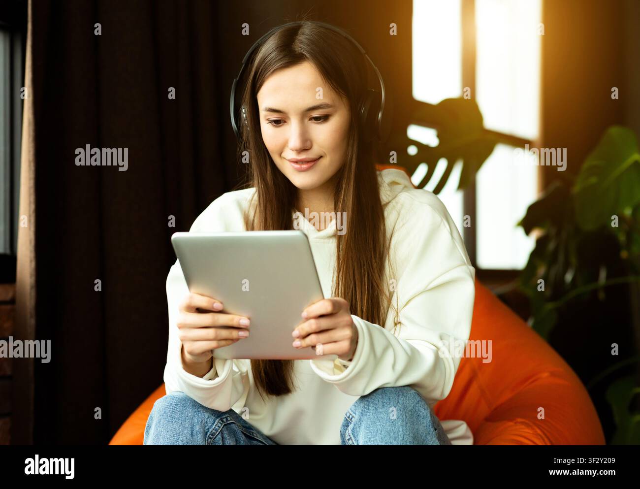 La ragazza moderna con tablet e cuffie funziona nella sedia a sacco Foto Stock