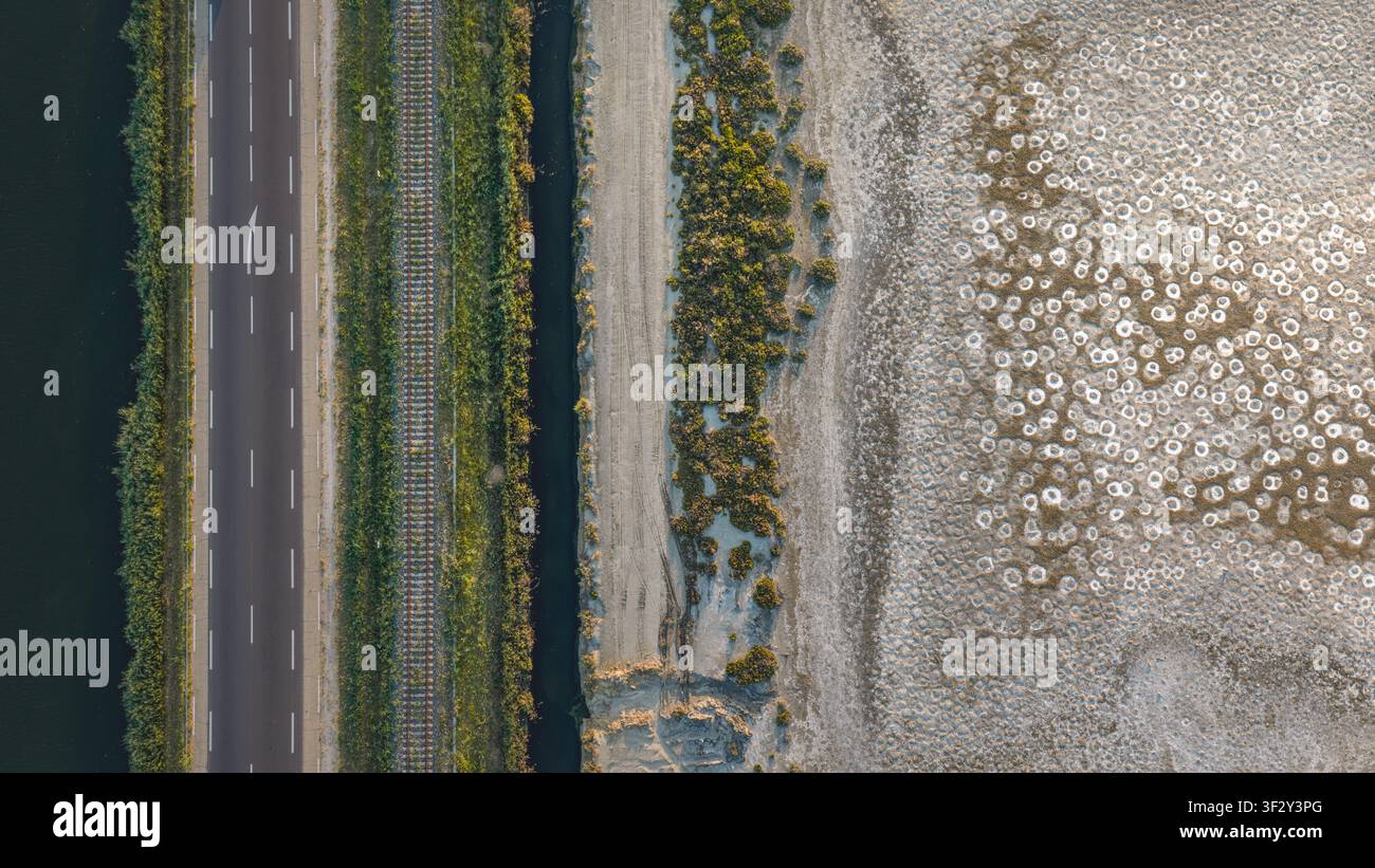 Vista aerea di un'infrastruttura di trasporto parallela che include una strada e binari ferroviari accanto a una palude salata con schemi minerali bianchi astratti sul g Foto Stock
