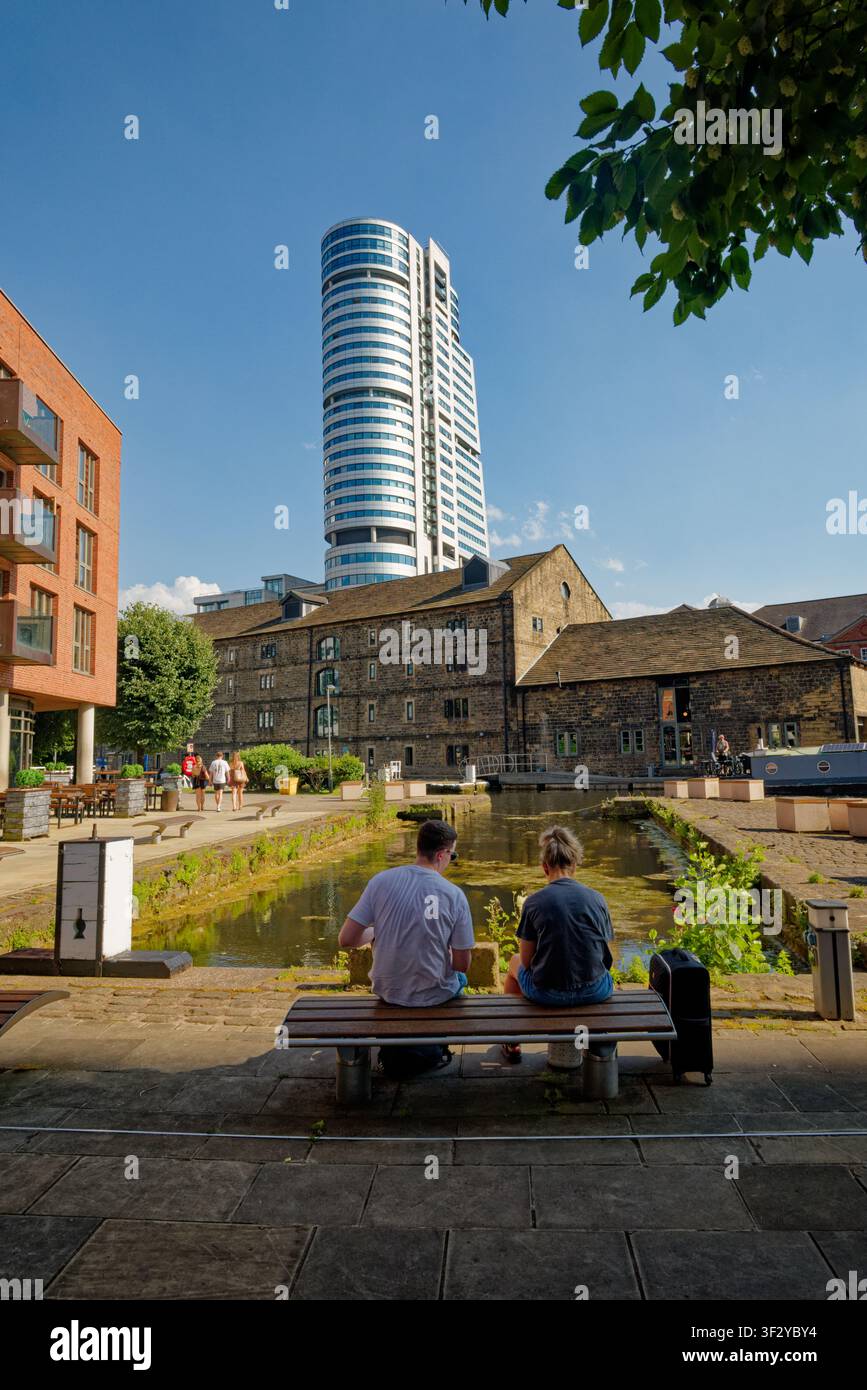 Una coppia (uomo e donna) è seduta su una panchina accanto a un bacino canale con un alto edificio per uffici sullo sfondo, Granary Wharf, Leeds, West Yorkshire. Foto Stock