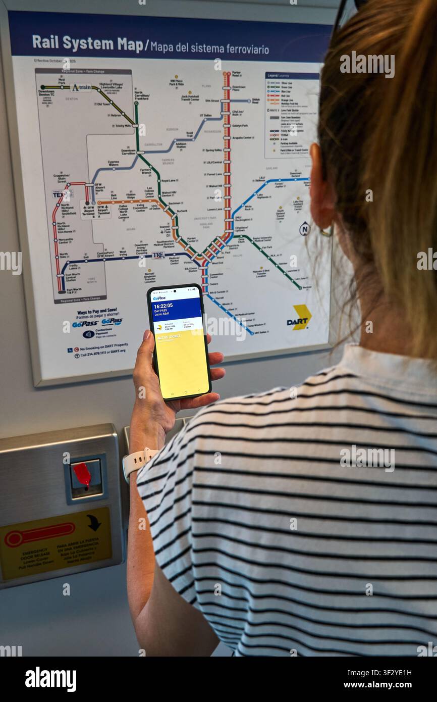 DALLAS, TX, USA - 11 LUGLIO 2026: Una donna tiene uno smartphone che mostra un'app di trasporto davanti a una mappa del sistema ferroviario DART. DART è un importante sistema di trasporto pubblico Foto Stock