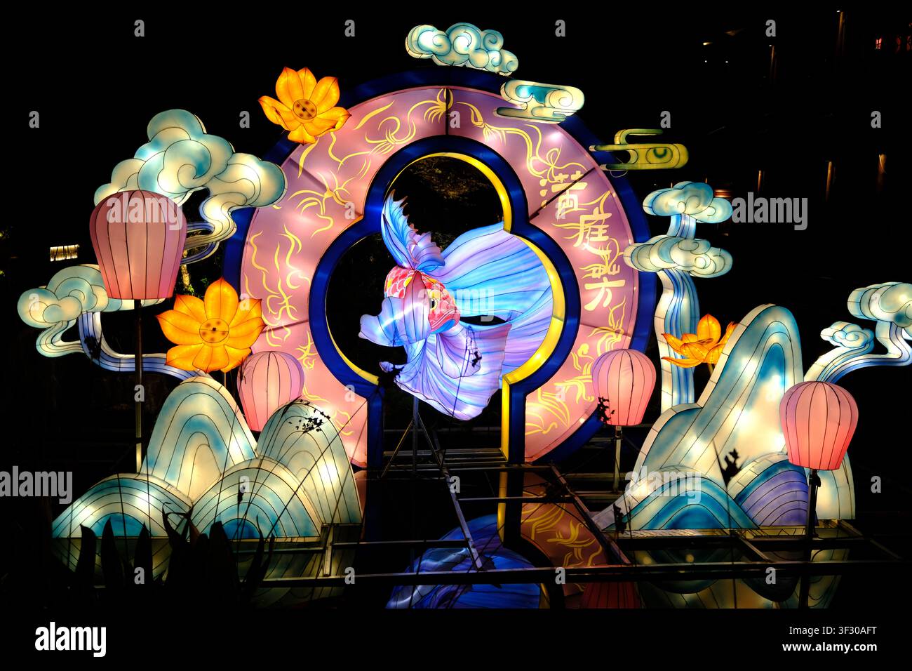 Esposizione di lanterne colorate notturne con una ballerina luminosa, lanterne rosa, fiori, nuvole e onde in una scena festiva all'aperto con sfondo scuro. Foto Stock