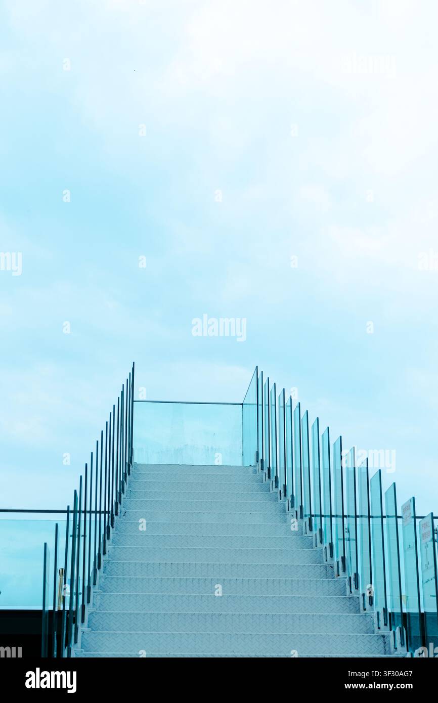 Scalinata esterna bianca minimale con ringhiere in vetro che porta verso l'alto in un cielo blu morbido, creando un aspetto moderno e pulito con spazio per il testo e una sensazione Foto Stock
