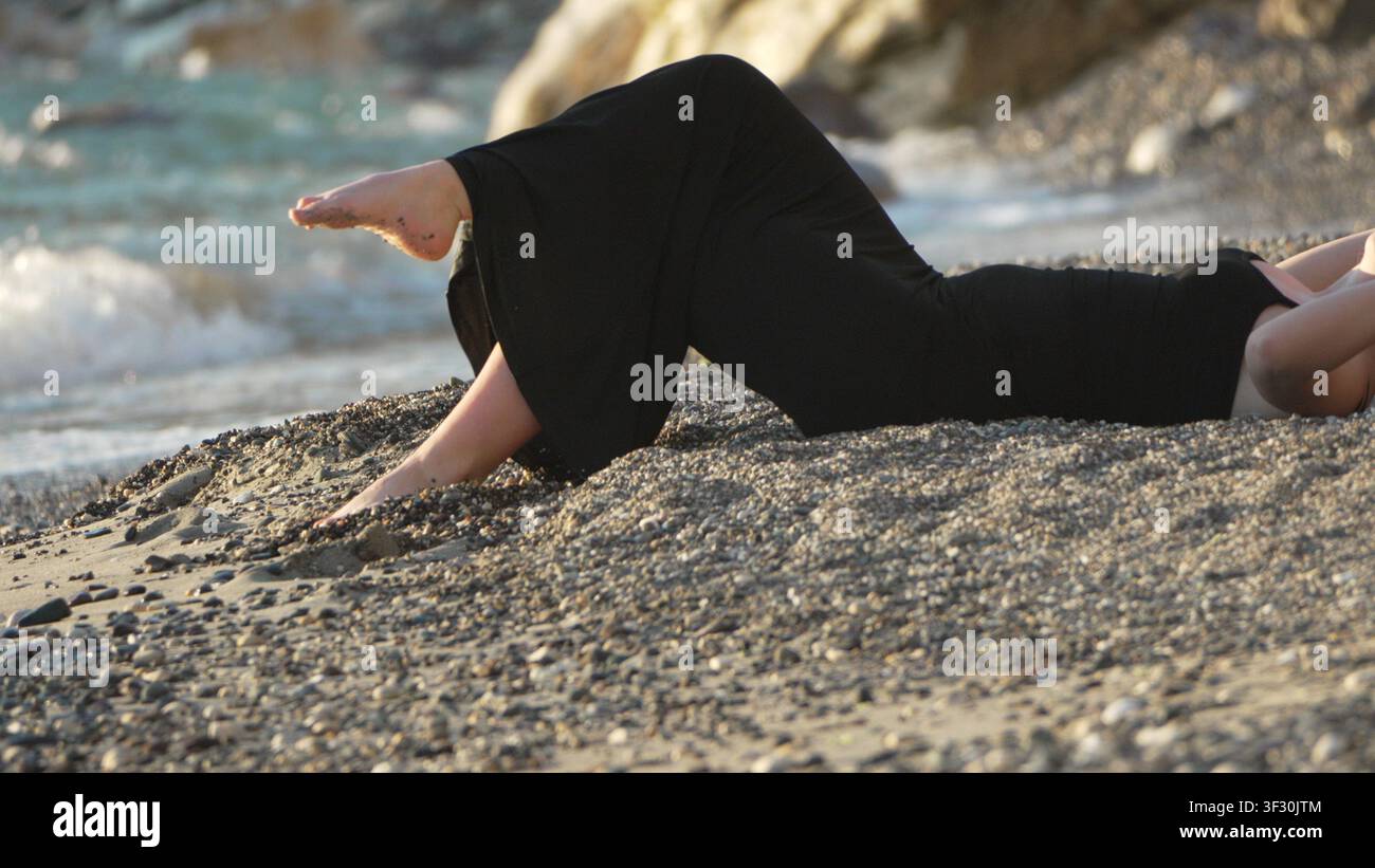 Vestito da spiaggia da donna. Una giovane donna in un elegante maxivestito nero che si rilassa su una bellissima spiaggia di ciottoli al tramonto Foto Stock