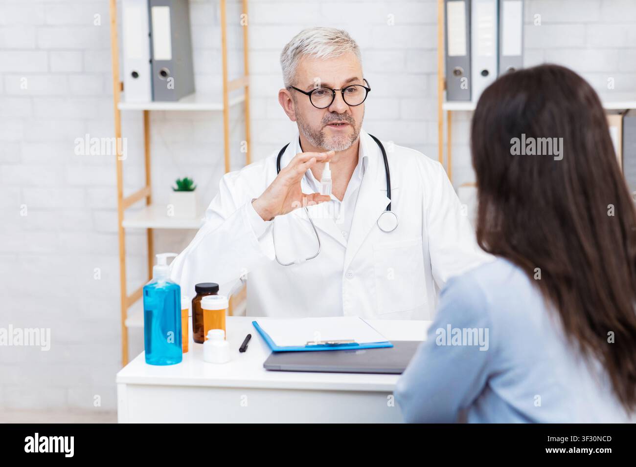 Serio medico dell'anziano in cappotto bianco e gli spettacoli dei vetri flacone a signora paziente al tavolo con vasi di pillole Foto Stock