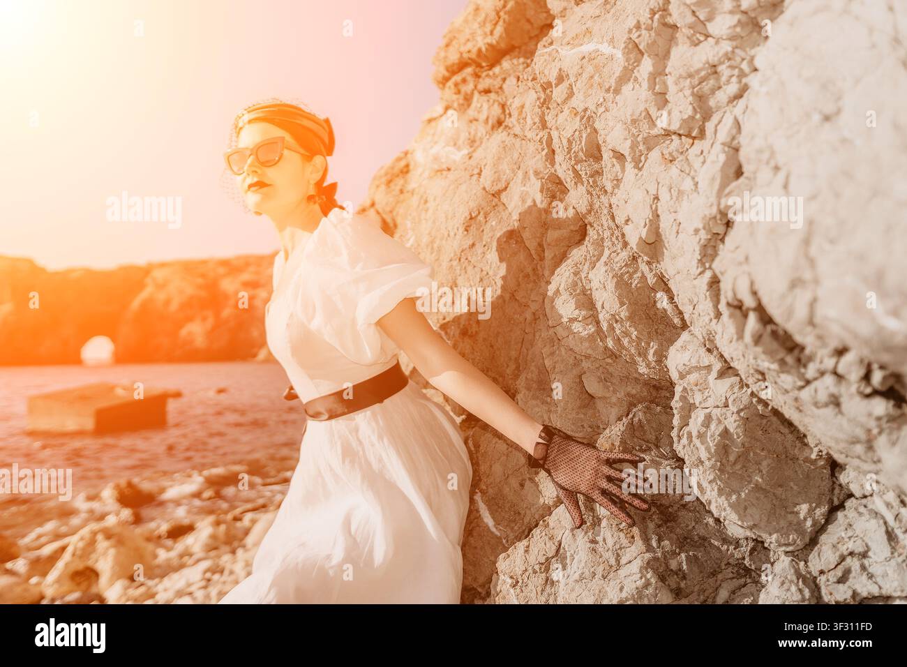 Donna vintage, signora costiera in abito bianco appoggiata sulle rocce durante il tramonto, catturando una nostalgia estiva onirica e vibrazioni vacanziere Foto Stock