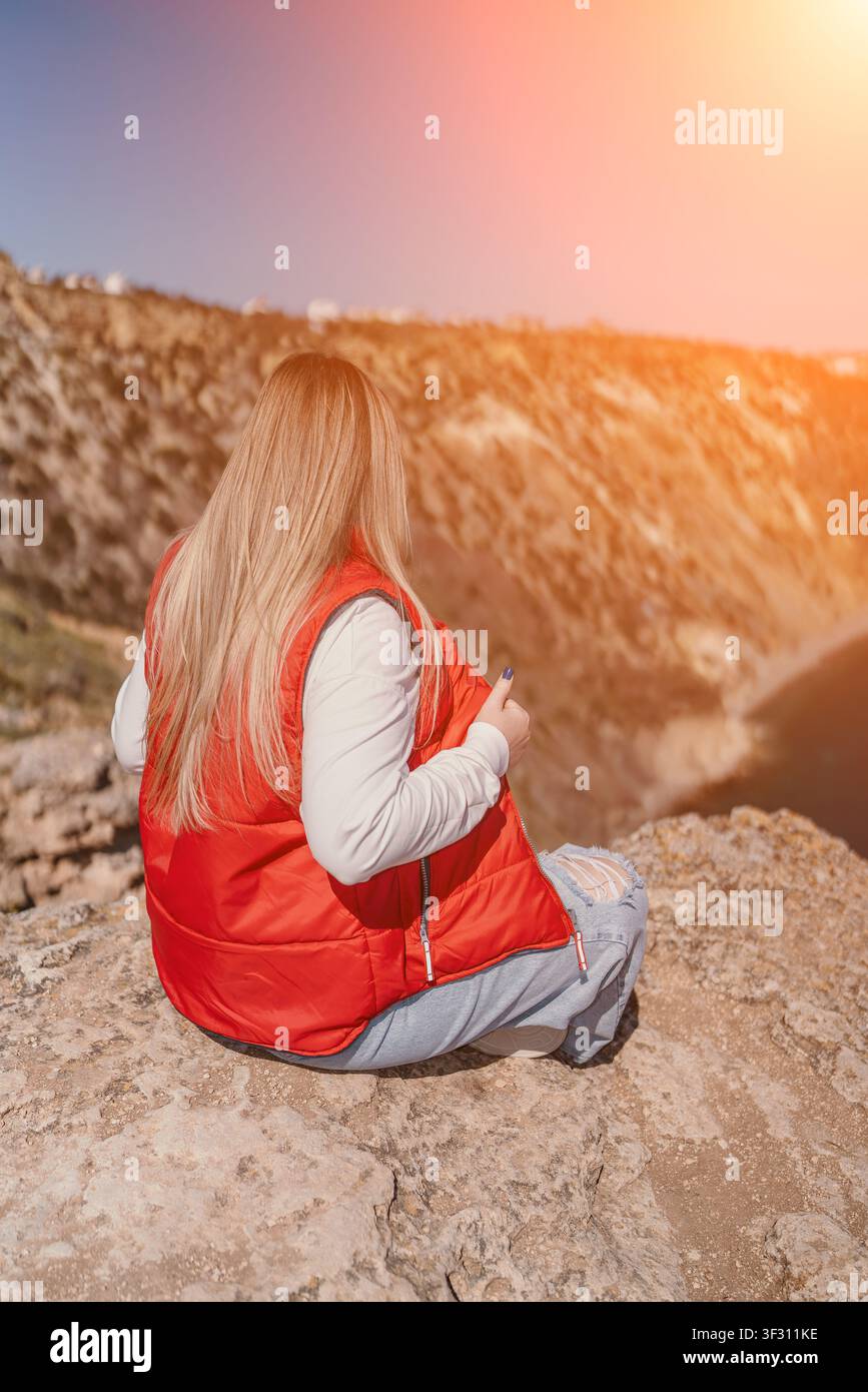 Donna sulla scogliera nella natura, seduta su un bordo roccioso che si affaccia sul mare durante una bellissima avventura di viaggio al tramonto dorato Foto Stock