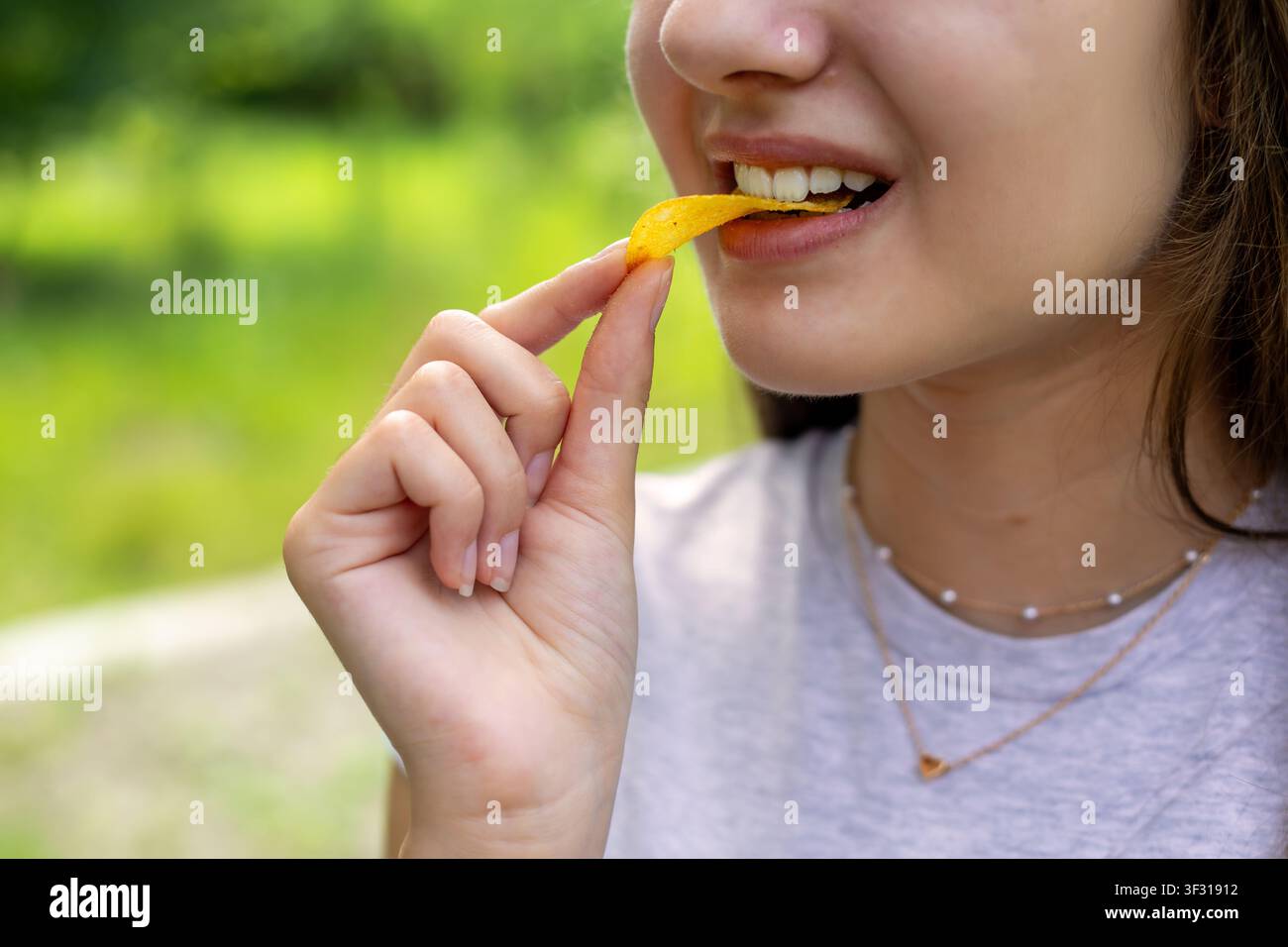 Primo piano di una donna che mangia una patatina fuori porta. Primo piano di una giovane donna che morde una patatina dorata all'aperto su uno sfondo verde sfocato. Foto Stock
