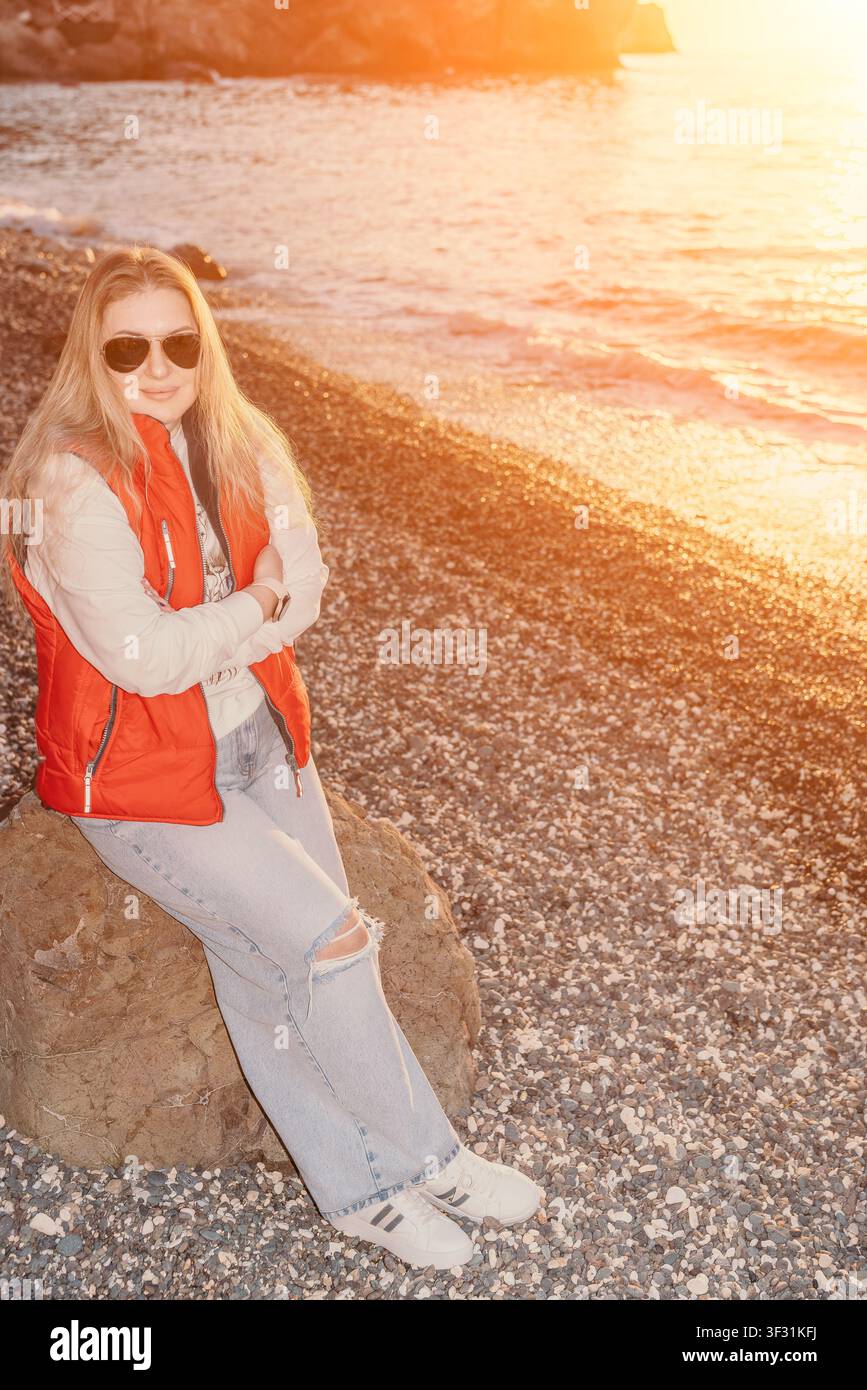 Donna seduta su una roccia sulla spiaggia al tramonto, indossa un giubbotto rosso sulla riva durante una bella ora dorata Foto Stock