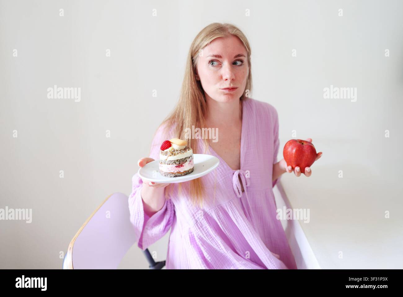 Giovane donna che sceglie tra una mela fresca e una torta deliziosa, illustrando un'alimentazione sana, l'equilibrio nutrizionale e le scelte alimentari quotidiane. Foto Stock