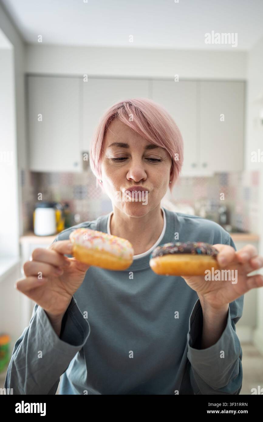 Donna matura pensierosa che mantiene una dieta e un peso sani, tiene dei ciambellini glassati decidendo di mangiare Foto Stock