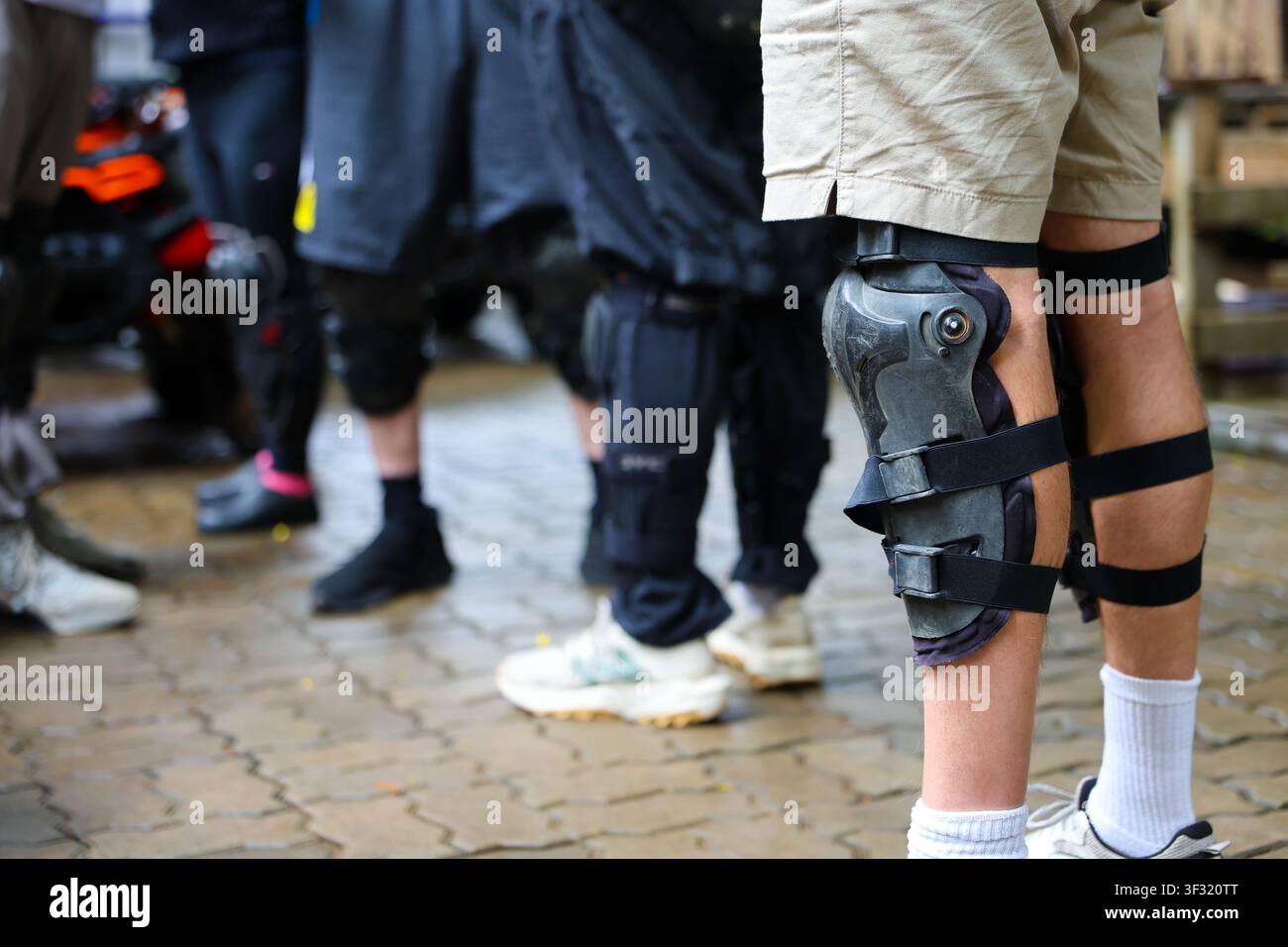 Un ginocchio protetto su una persona in piedi all'aperto con pantaloncini e calzini sportivi, un gruppo di persone e un marciapiede bagnato sullo sfondo che trasmettono sostegno attivo e R Foto Stock