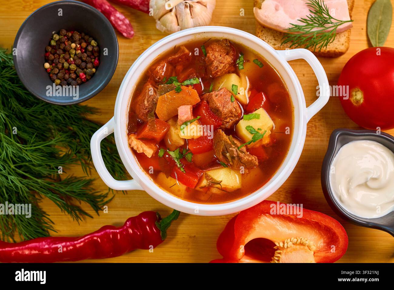 ciotola di tradizionale zuppa bograch ucraina o ungherese, presentata su una tavola di legno con un contorno di panna acida, un classico sandwich al salo, aneto fresco, peperoncini Foto Stock