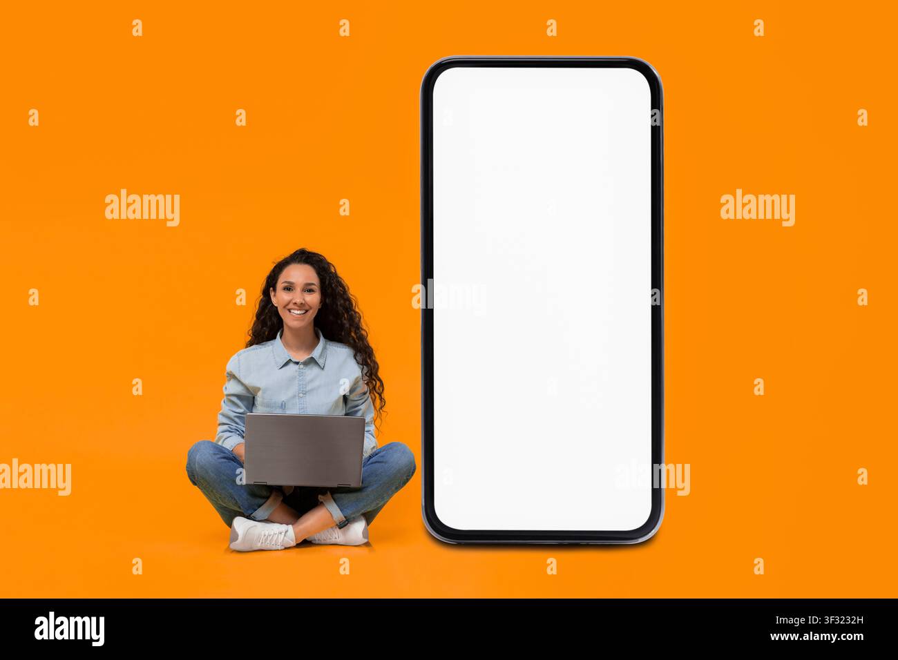Donna che utilizza un laptop seduta sul pavimento accanto a uno schermo grande e vuoto per smartphone Foto Stock