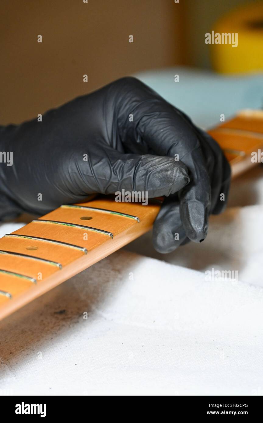 Primo piano di un tecnico chitarrista o liutista che indossa un guanto in nitrile nero e tiene un collo di chitarra elettrica in acero staccato con tasti durante la pulizia, la lucidatura e i lavori di regolazione. Manutenzione professionale degli strumenti, artigianato liutario e processo di officina. Ideale per concetti di servizio musicale, officina di riparazione, cura della chitarra e artigianato. Foto Stock