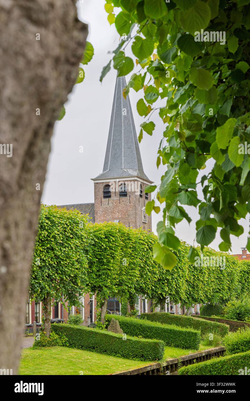Una torre di chiesa storica in mattoni si erge sopra una fila di tigli potati e siepi formali lungo un tranquillo canale a IJlst, Friesland, Paesi Bassi, evidenziando Foto Stock