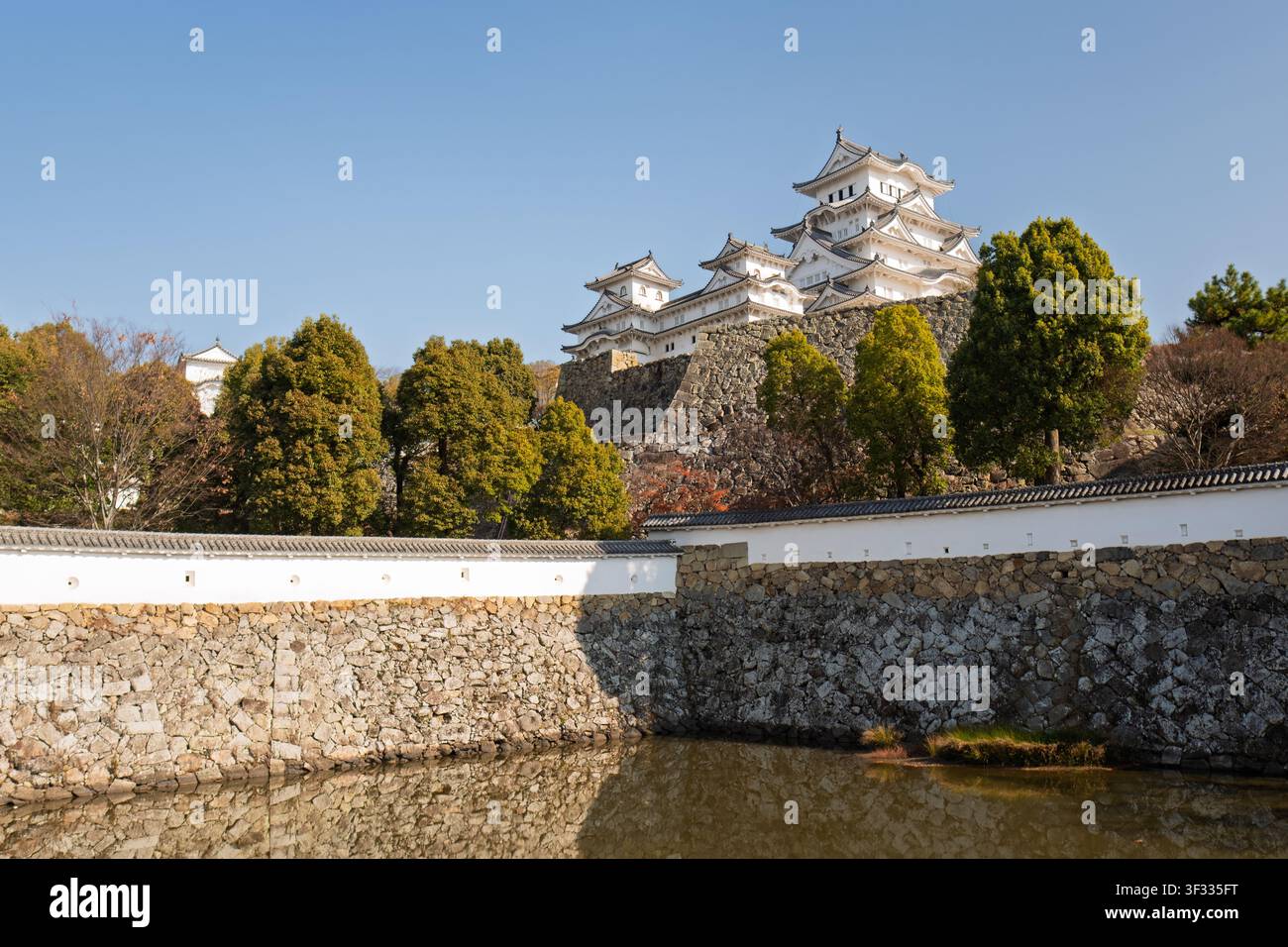 Vista grandangolare del mastio del castello di Himeji che si erge dietro alberi verdi di un parco e un fossato d'acqua protettivo in pietra, Giappone Foto Stock