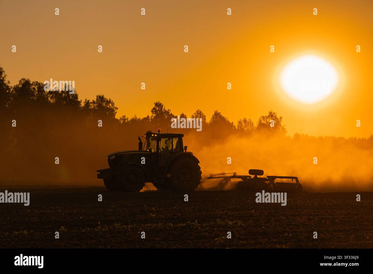 Trattore agricolo moderno che coltiva un campo al tramonto dorato con polvere Foto Stock
