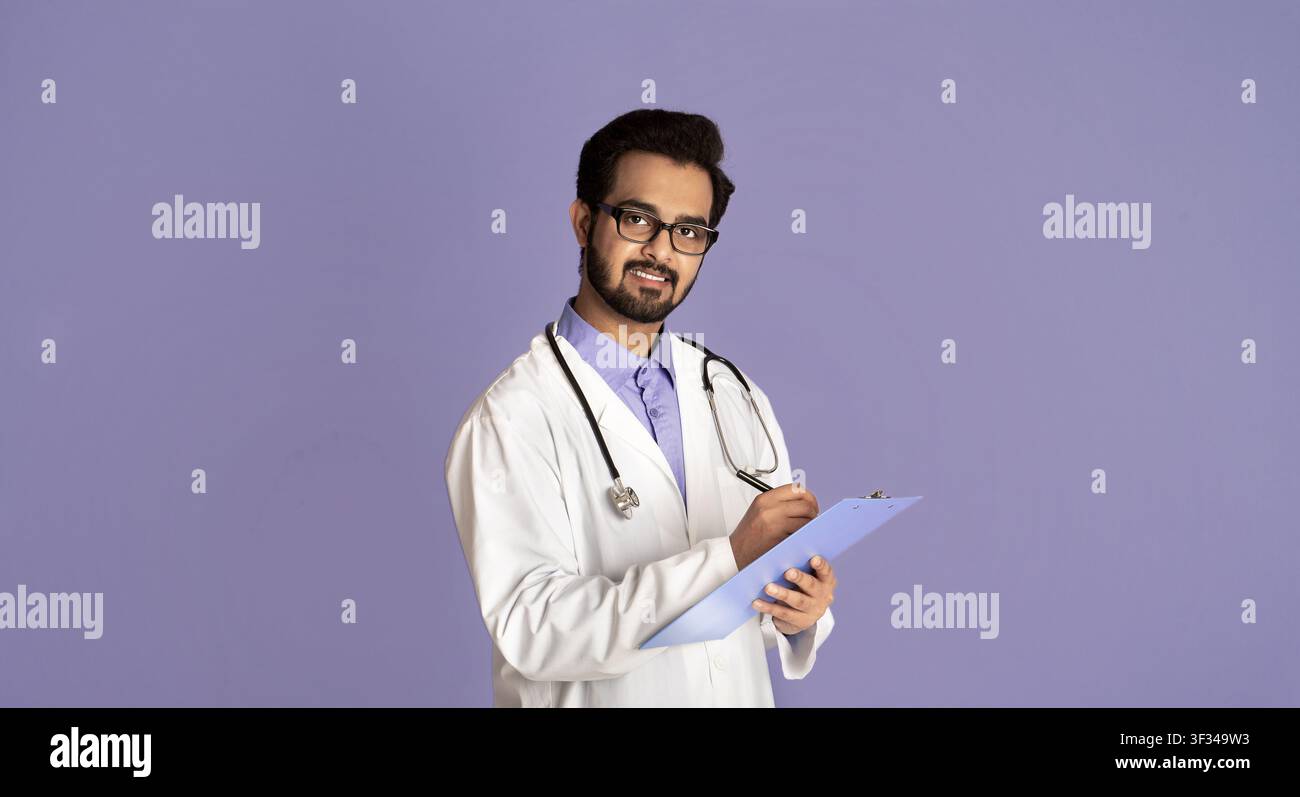 Ritratto di giovane medico indiano maschio che scrive sulla clipboard sopra sfondo lilla Foto Stock