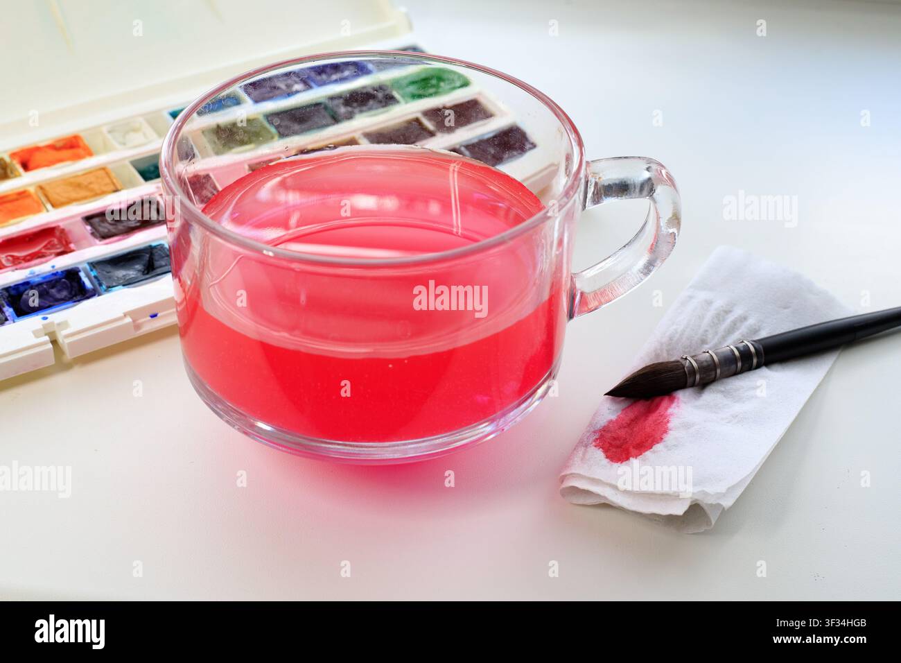 Spazio di lavoro di un artista ad acquerello con un bicchiere d'acqua, un pennello, un tovagliolo macchiato e una tavolozza con colori ad acquerello su un tavolo bianco. Processo creativo Foto Stock