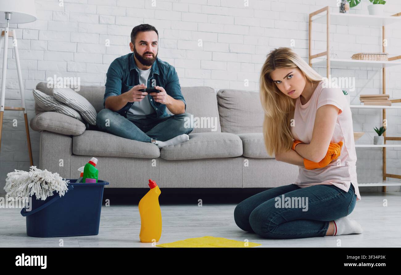 Indifferenza e pigrizia. L'uomo gioca in videogiochi seduti sul divano con joystick, donna offesa ha attraversato le braccia sul petto e lavano il pavimento Foto Stock