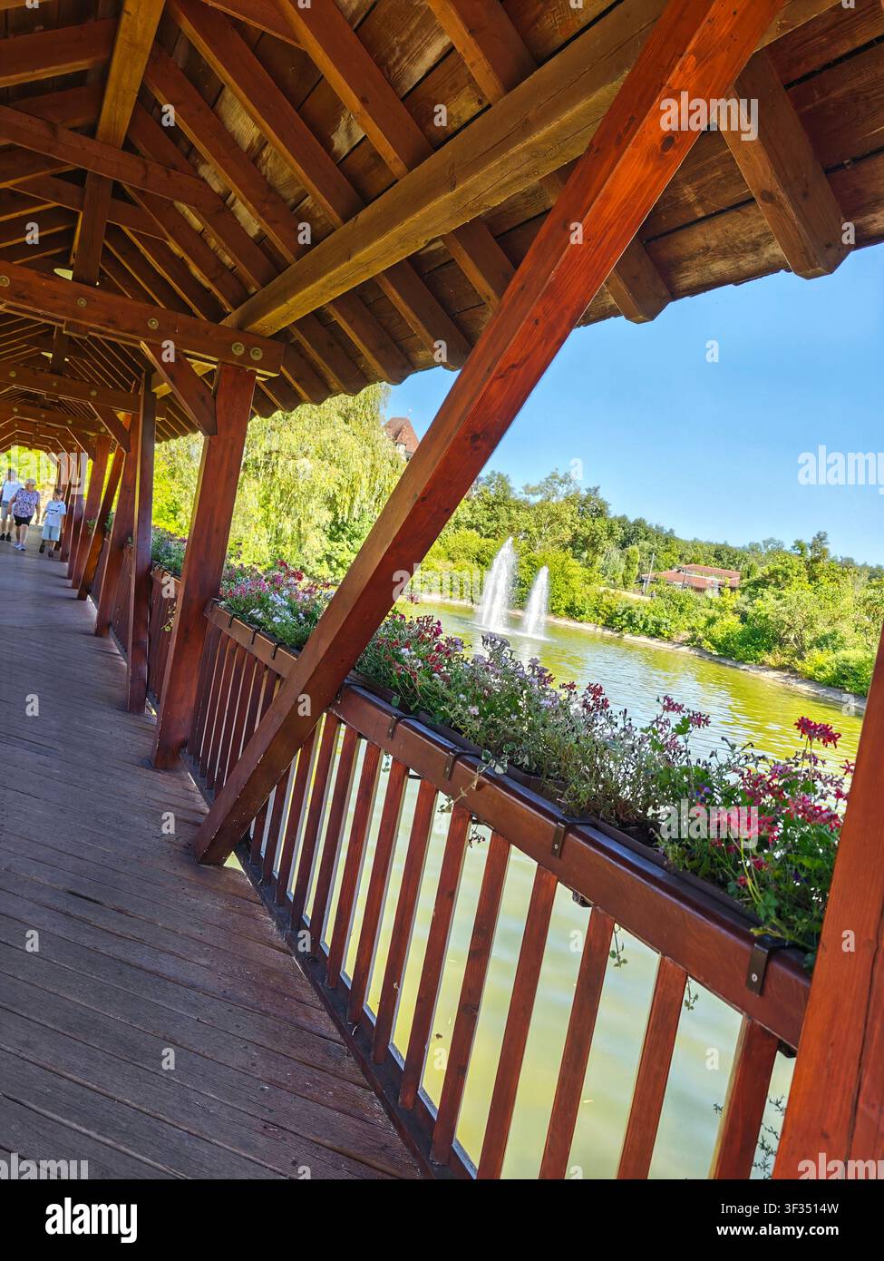 Ponte in legno coperto tradizionale con fioriere che si affaccia su uno stagno con fontane all'Erlebnispark Tripsdrill, Cleebronn, Baden-Württemberg, Germania - Immagine stock catturata con smartphone