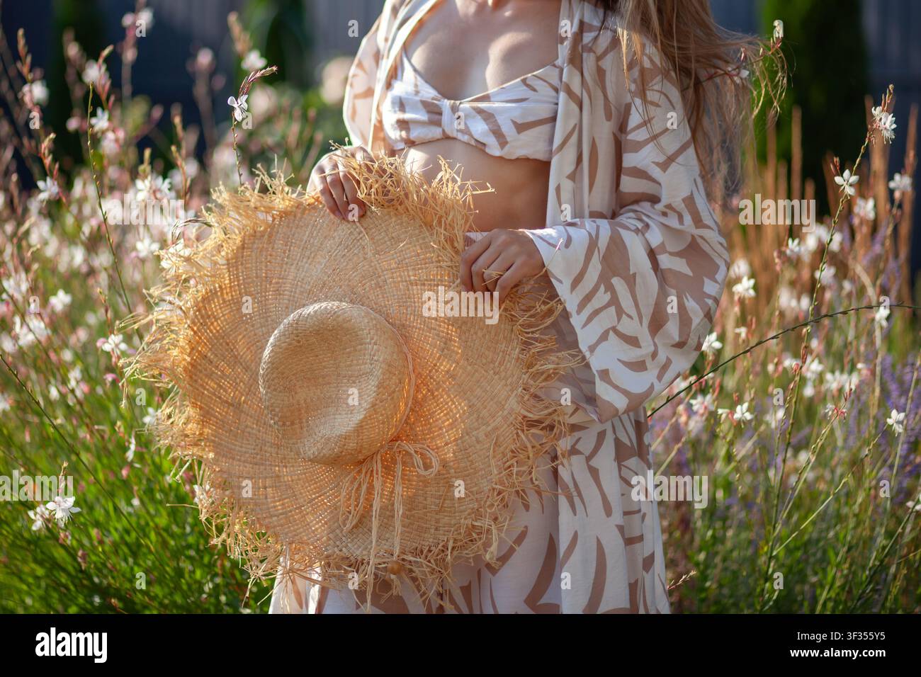 Donna elegante tiene un cappello di paglia, indossa un set alla moda di reggiseno e pantaloni da accappatoio in un giardino estivo. Primo piano del bordo sfilacciato del cappello Foto Stock