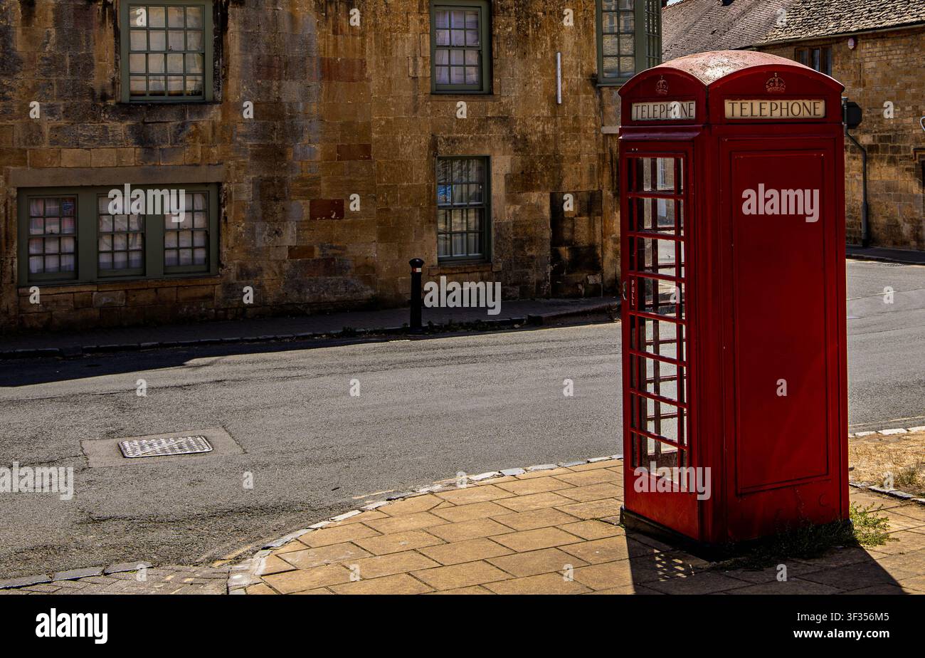 Iconica cabina telefonica rossa in piedi su un marciapiede lastricato accanto a una strada asfaltata e a un edificio storico in pietra a Chipping Campden, Inghilterra Foto Stock