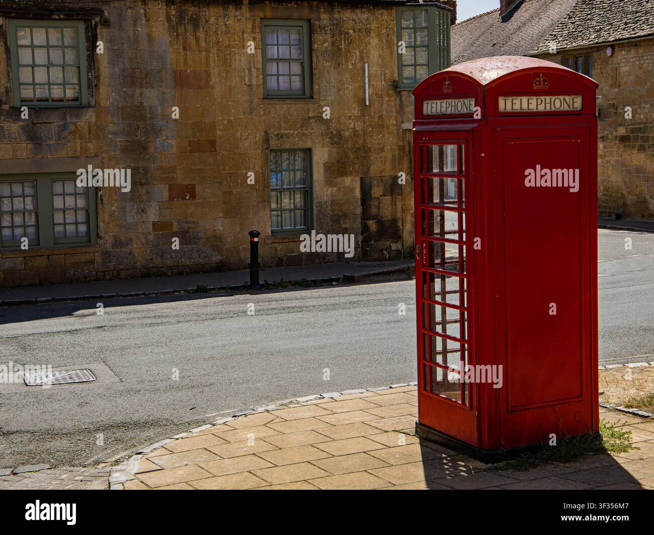 Iconica cabina telefonica rossa in piedi su un marciapiede lastricato accanto a una strada asfaltata e a un edificio storico in pietra a Chipping Campden, Inghilterra Foto Stock